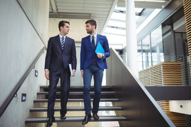 Two businessmen in suits are walking downstairs in a modern office building. They appear to be engaged in a discussion, possibly heading to a meeting or collaborating on a project. This image can be used for business-related content, corporate websites, teamwork and collaboration themes, or professional services marketing.