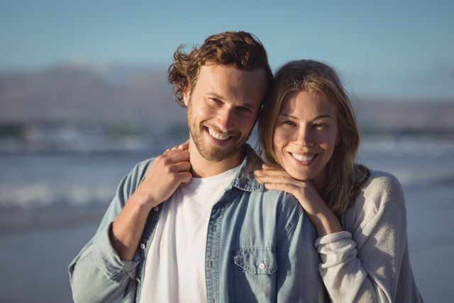 This image captures a happy couple standing close together at the beach on a sunny day. They are both smiling, showcasing their joy and affection for each other. Ideal for use in advertisements, travel brochures, lifestyle blogs, and social media posts promoting love, relationships, and outdoor activities.