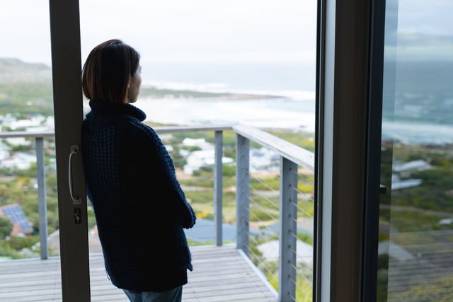 Asian woman leaning on window, gazing at scenic landscape from balcony. Ideal for themes of solitude, reflection, relaxation, and nature. Suitable for articles on mental health, personal growth, and peaceful living.