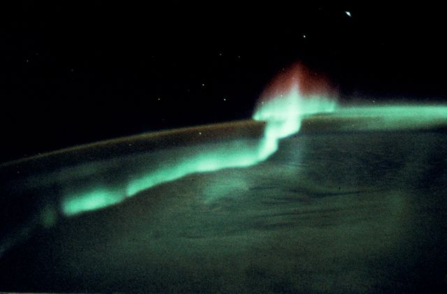 Astronaut Robert F. Overmyer captures stunning auroras in the Southern Hemisphere during a 1985 space mission. The image features blue-green auroras with tall red rays over Earth's horizon, surrounded by moonlit clouds. Dr. Don L. Lind, a mission specialist, pinpointed the location as midway between Australia and Antarctica. Ideal for educational material on space and atmospheric phenomena, science presentations, or backgrounds highlighting natural marvels.