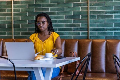 African american woman working on laptop in modern cafe drinking coffee with croissant