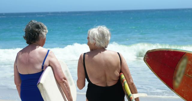 Two senior women are standing on a sunny beach holding surfboards, facing the ocean with waves breaking in the background. Both are dressed in swimsuits and ready to enjoy surfing. This image can be used for promoting healthy and active lifestyles among seniors, vacation advertisements, retirement communities, and more.