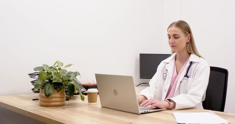Doctor typing on laptop in minimalist clinic office wearing white coat and stethoscope