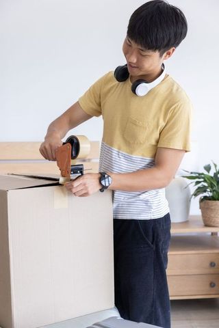 Young asian man sealing moving box with tape dispenser in minimalist bright home interior