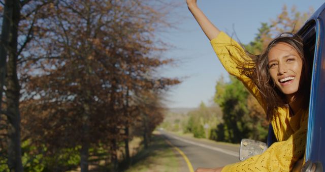 Young woman smiling and enjoying a road trip with her arm out of a car window; perfect for use in travel blogs, car advertisements, adventure-themed campaigns, promoting road safety, or freedom lifestyle concepts.