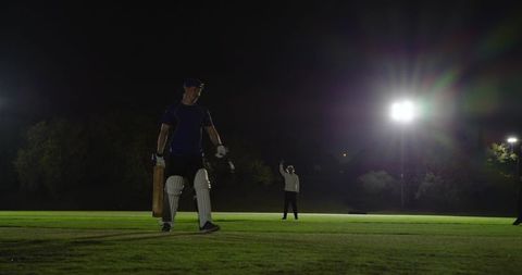 Night cricket batsman walking off under floodlights while umpire signaling dismissal
