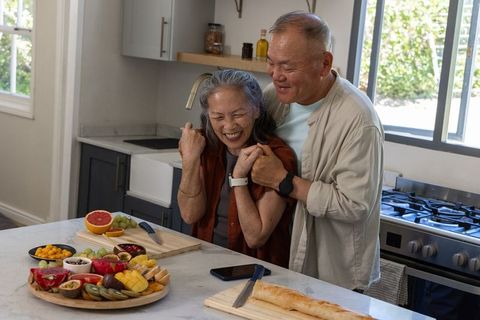 Senior asian couple laughing and hugging in bright kitchen with fruit platter and baguette