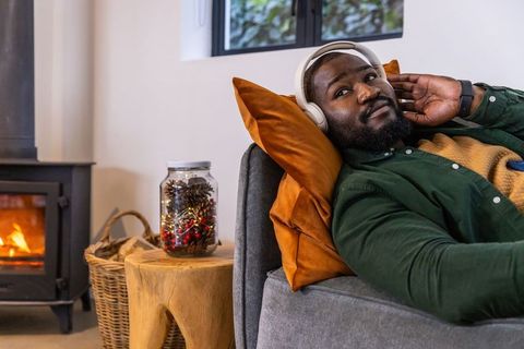 African american man relaxing on sofa with headphones by cozy wood stove and seasonal decor