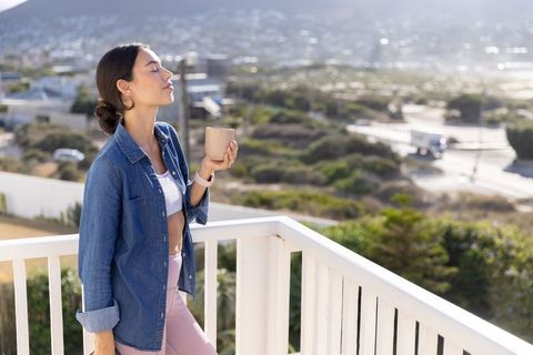 Woman inhaling fresh air on balcony holding mug overlooking coastal hillside morning