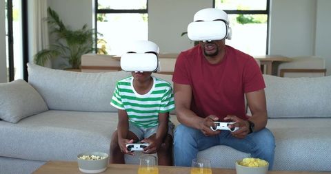 African American Father and Son Playing VR Games on Couch with Headsets and Controllers