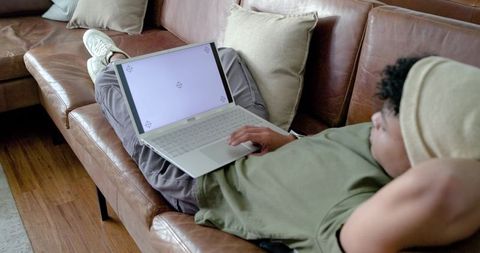 Adult man reclining on leather sofa using laptop wearing bucket hat relaxing at home
