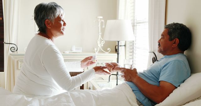 A middle-aged African American woman is sitting by the bedside, holding hands with a man who appears to be a patient, with copy space. Their interaction suggests a moment of care and support, within a home or healthcare setting.