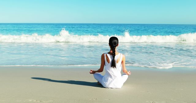 A woman wearing a white outfit is meditating on a sandy beach, facing the ocean waves with a calm and peaceful expression. Ideal for wellness and relaxation themes, mindfulness, vacation and travel promotion, mental health awareness, and lifestyle blogs. This image conveys a sense of tranquility and serene connection with nature.