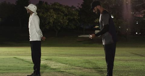 Two men standing on twilight grass field under floodlight wearing bucket hat and cap