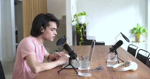 Young man in twenties wearing pink top recording podcast at home studio desk with laptop