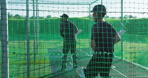 Teenager practicing cricket batting in green net cage with coach during outdoor training