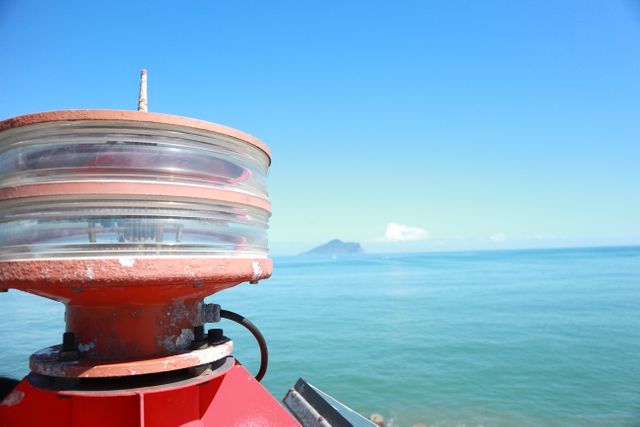 Close-up view of a maritime light with the ocean and an island in the background on a clear, sunny day. Ideal for use in travel brochures, navigation themed content, or any artwork pertaining to maritime adventures or coastal living.