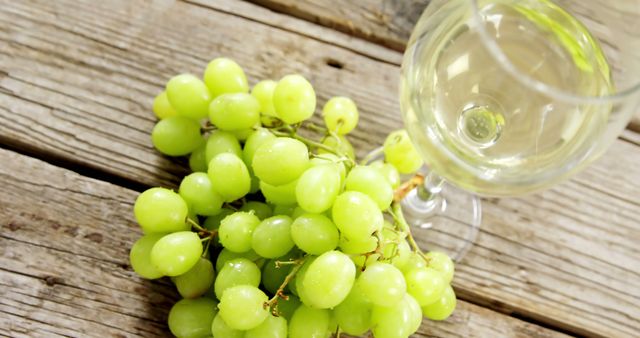 A bunch of green grapes is placed next to a glass of white wine on a rustic wooden table, with copy space. Grapes are often associated with wine production, and this setting suggests a tasting or the enjoyment of fresh produce and wine.