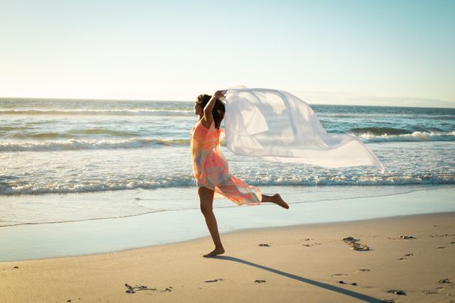 Biracial woman joyfully dancing on beach while holding a flowing scarf. Ideal for themes of freedom, happiness, and leisure. Perfect for travel brochures, lifestyle blogs, and advertisements promoting beach vacations and outdoor activities.