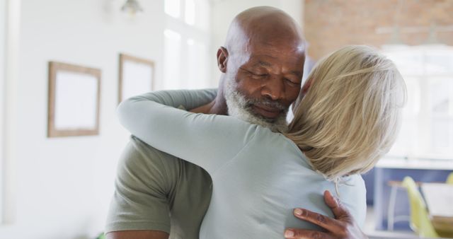Senior couple seen hugging each other closely in a bright, sunlit room. Ideal for use in topics related to relationships, senior living, love, and support among elderly couples. Can be utilized in promotional materials for retirement homes, healthcare, or articles on aging and family bonds.