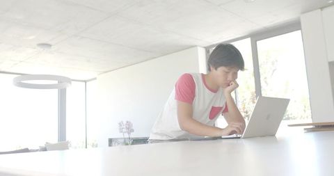 Leaning asian man working on laptop at modern bright kitchen island with minimal open-plan interior