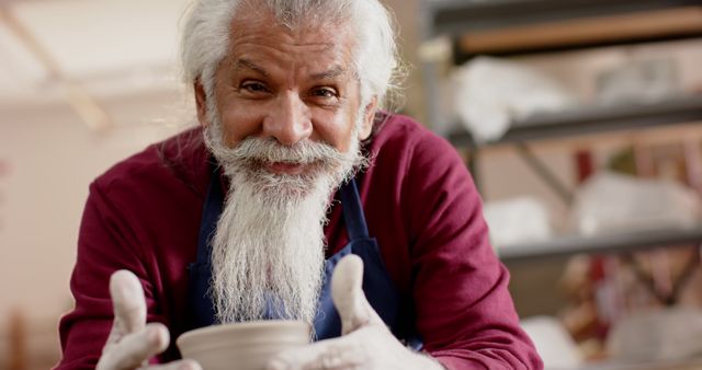 An elderly man with a friendly expression and gray beard works intently on a clay sculpture in an art studio. The focus is on his detailed, skilled hands and content face, highlighting the joy he finds in his craft. This image is perfect for promoting craftsmanship, hobbies in retirement, and inspirational stories about finding joy in artistic pursuits.