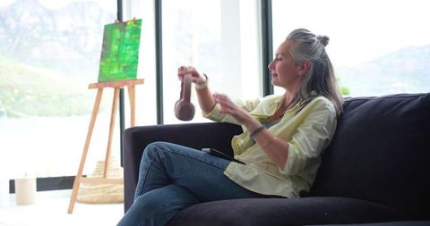 Senior woman relaxing on sofa holding headphones and checking tablet with lake view