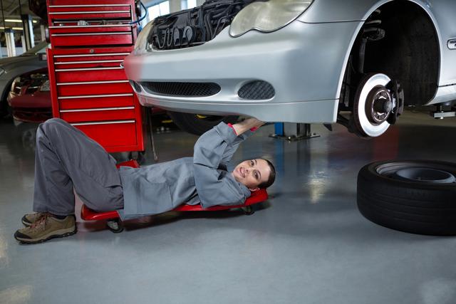 Female mechanic lying on a creeper and repairing a car in an auto garage. Ideal for use in articles or advertisements about women in skilled trades, automotive repair services, and gender diversity in the workplace. Can also be used for educational materials on car maintenance and vocational training.