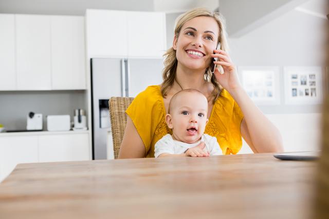 This image shows a joyful mother multitasking by holding her baby while talking on the phone in a bright, modern kitchen. Use this image for content related to parenting, family life, motherhood, work-life balance, modern technology, or home living. It can be ideal for blogs, articles, advertisements, and social media posts focused on parenting tips, family happiness, or domestic life.