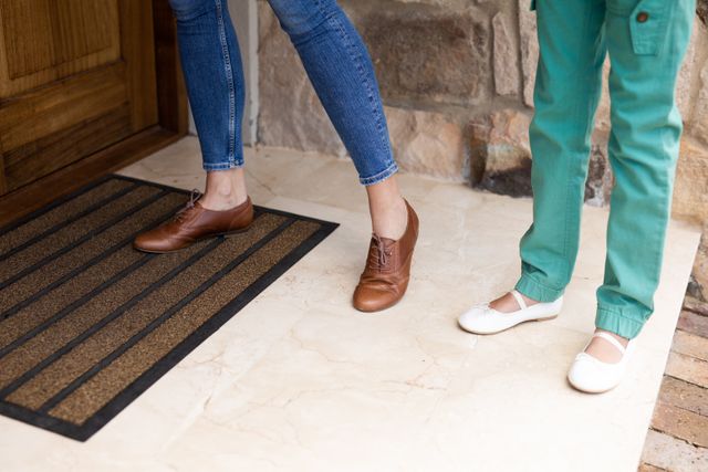 Caucasian mother and daughter's feet outside the front door about to enter their house. the mother's other foot is stepping on the doormat.
