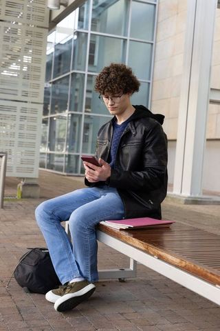 Teenage student sitting on bench using smartphone near modern glass building with backpack and folde