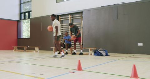 African american teammates practicing basketball dribbling through cones in gym training