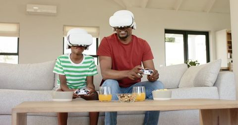 African American father and son playing VR games at home wearing headsets and holding controllers