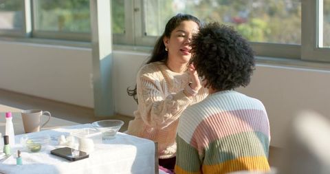 Sunlit multiracial friends applying makeup together by window for relaxed beauty routine