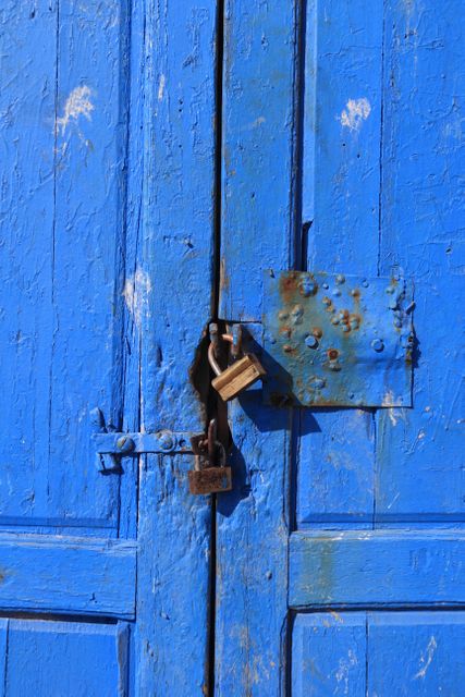 Vivid blue-painted wooden door with visible weathering and signs of aging, secured with strong padlock. Perfect for themes showcasing security, rustic charm, vintage look, and old architecture. Ideal for use in blog posts, editorials, and home decor articles focusing on historical buildings and artistry in decay.