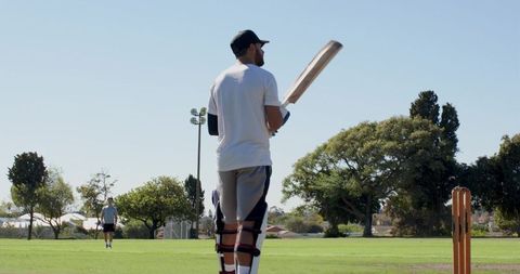 Cricket batter standing holding wooden bat preparing to face bowler on grassy pitch