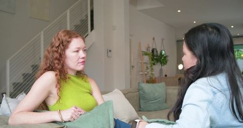 Redhead and asian women sitting on sofa talking and listening in modern cozy living room
