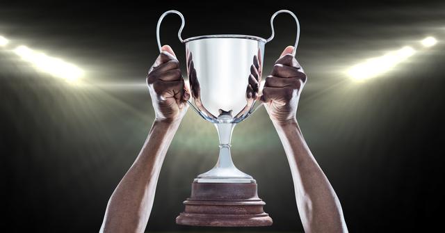 Hands of an athlete lifting a trophy in front of bright spotlights, capturing the moment of victory and celebration. Suitable for promoting sports events, achievement themes, motivational content, and success-driven campaigns focused on triumph and hard work.
