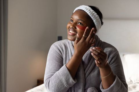 African american woman applying red under-eye hydrogel patches smiling in cozy robe