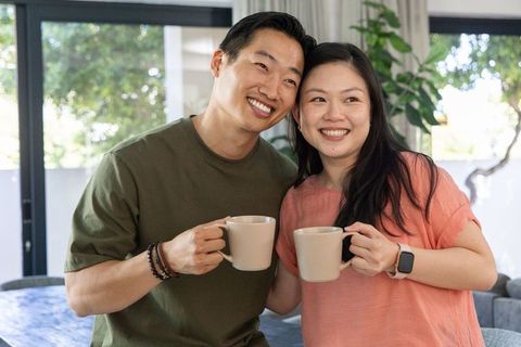 Asian couple smiling and holding coffee mugs in cozy sunlit home morning