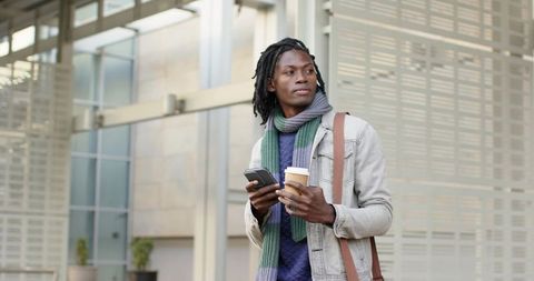 African American commuter holding smartphone and coffee, wearing scarf and denim jacket