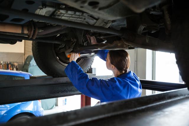 Female mechanic in blue uniform servicing car undercarriage at repair garage. Ideal for illustrating automotive services, gender diversity in technical professions, and professional car maintenance.