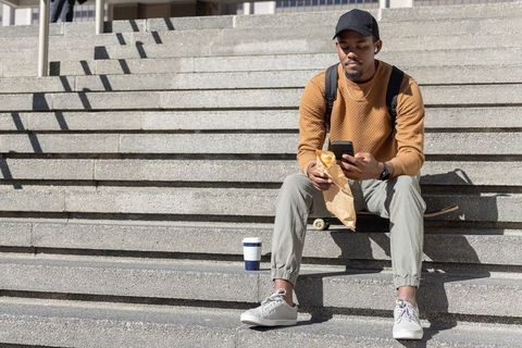 African american man sitting on concrete steps checking phone with skateboard and coffee