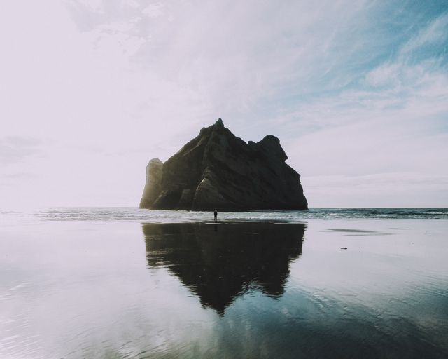 This image captures a solitary rock formation standing tall on a tranquil beach with its reflection mirrored on the shoreline. Sparse human presence emphasizes the feeling of solitude and connection with nature. Perfect for designs or articles that celebrate natural beauty, solitude, or outdoor adventures. Can be used in travel brochures, meditation blogs, or environmental posters.