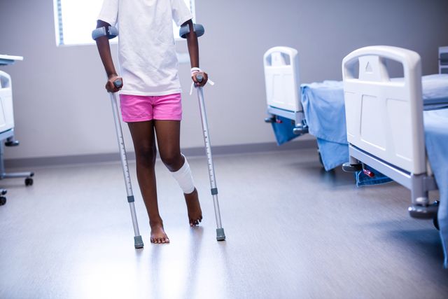 A young girl with a leg injury walking with crutches in a hospital ward, suggesting recovery and rehabilitation. Suitable for use in medical and healthcare contexts, especially those related to pediatric care, injury recovery, and hospital environments.