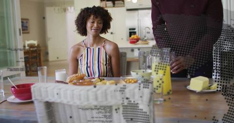 Curly-haired woman smiling at breakfast while partner pours orange juice in sunlit kitchen