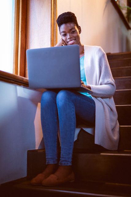 African American woman sitting on stairs at home, using laptop and smiling. Ideal for content related to remote work, home lifestyle, self care, and technology use in everyday life. Perfect for blogs, articles, and advertisements focusing on work-life balance, relaxation, and modern living.