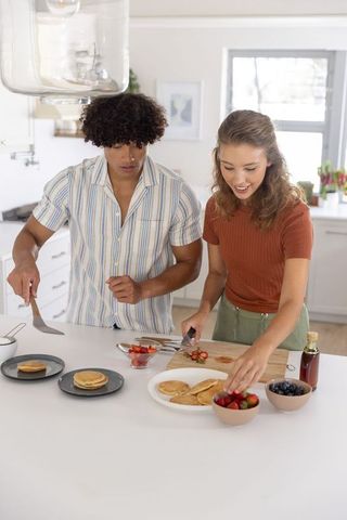 Diverse couple preparing breakfast in bright modern kitchen with pancakes and fresh berries