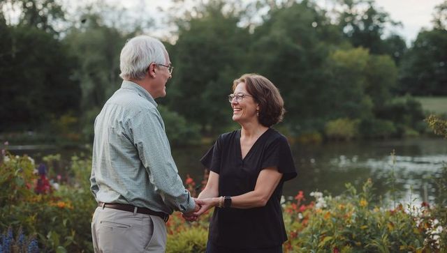 Senior couple holding hands and smiling by pond in garden park during golden hour romance