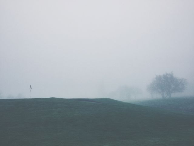 A serene golf course blanketed in heavy fog on a misty morning, creating a quiet and mysterious atmosphere. The scene is tranquil with limited visibility, adding an eerie yet peaceful mood. Trees and a flag are barely visible through the dense fog. This image can be used in articles or advertisements focusing on golf, nature, tranquility, or misty landscapes. It is a powerful visual for themes of solitude, calmness, and serene beauty.